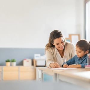 A professional female mentor helping a young child with a school assignment in a bright classroom